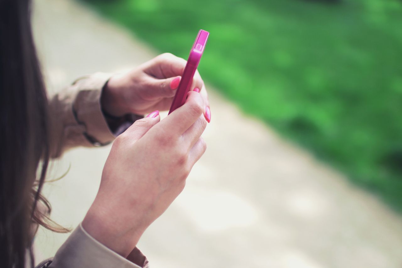 home-hero-bg Woman holding a pink smartphone with both hands in a casual outdoor setting.